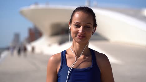 Young-woman-in-earphones-smiling-at-camera-on-street